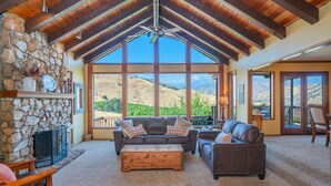 A spacious living room with a stone fireplace, large windows offering a view of rolling hills, and a ceiling fan. - Ranch Getaway in the foothills of the Sierras (Springville)