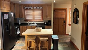 A kitchen with wooden cabinets, a black refrigerator, a wooden dining table with two stools, a window with blinds, and a door with a decorative mirror. - Deer Valley Townhouse, with New Private Hot Tub Just Steps Away from the Bus (Park City)