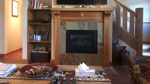 A living room with a fireplace, a flat-screen TV mounted above it, a bookshelf, and a coffee table with remote controls. - Deer Valley Townhouse, with New Private Hot Tub Just Steps Away from the Bus (Park City)