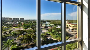 A view through a window of a parking lot with trees, a building with a green roof, and a distant view of a cityscape.