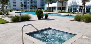 Zona de piscina de hotel con un jacuzzi, sillas de descanso y vista al edificio de fondo.