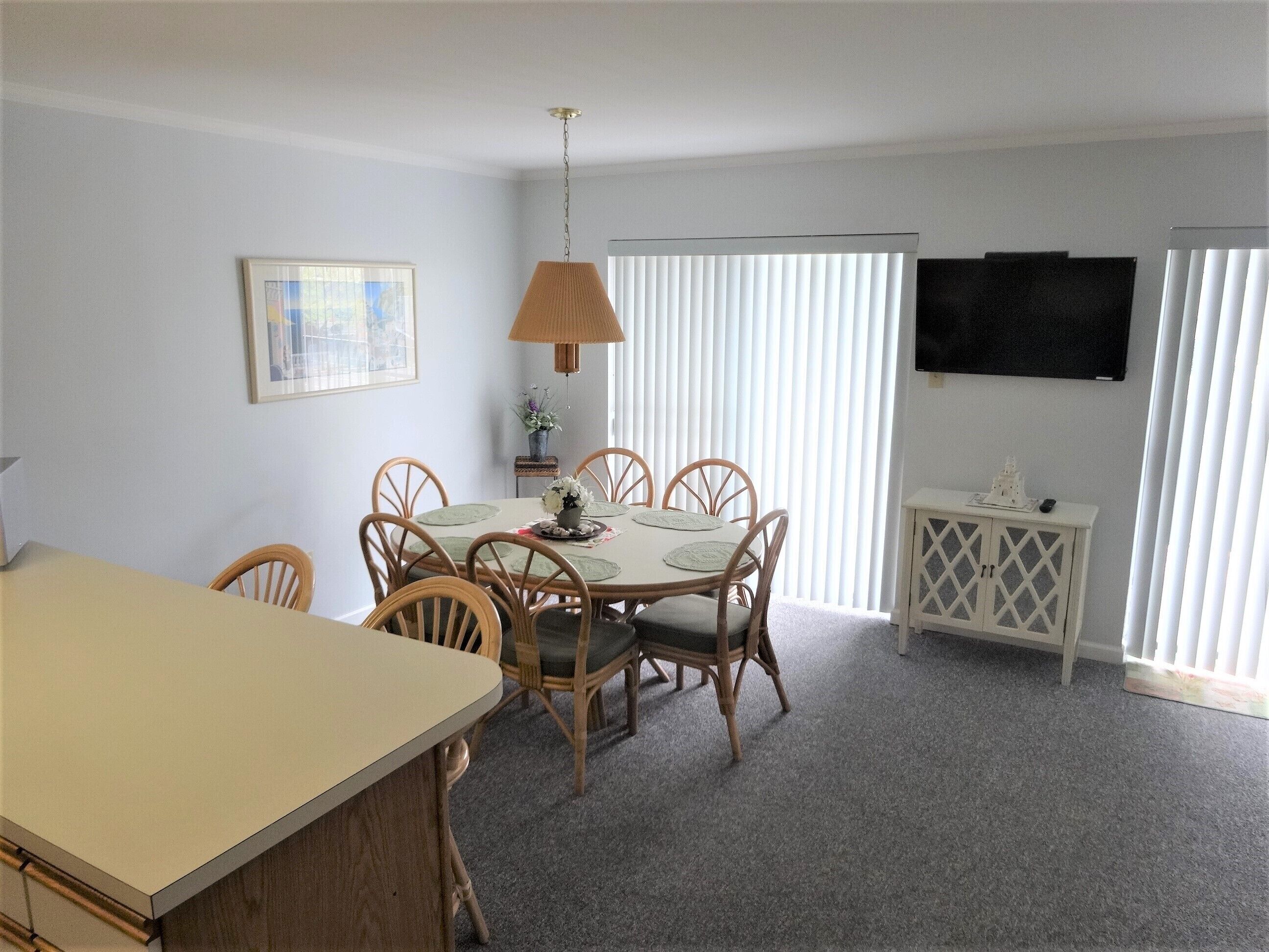 A dining area with a round table, six chairs, a hanging lamp, a small cabinet, and a television on the wall.