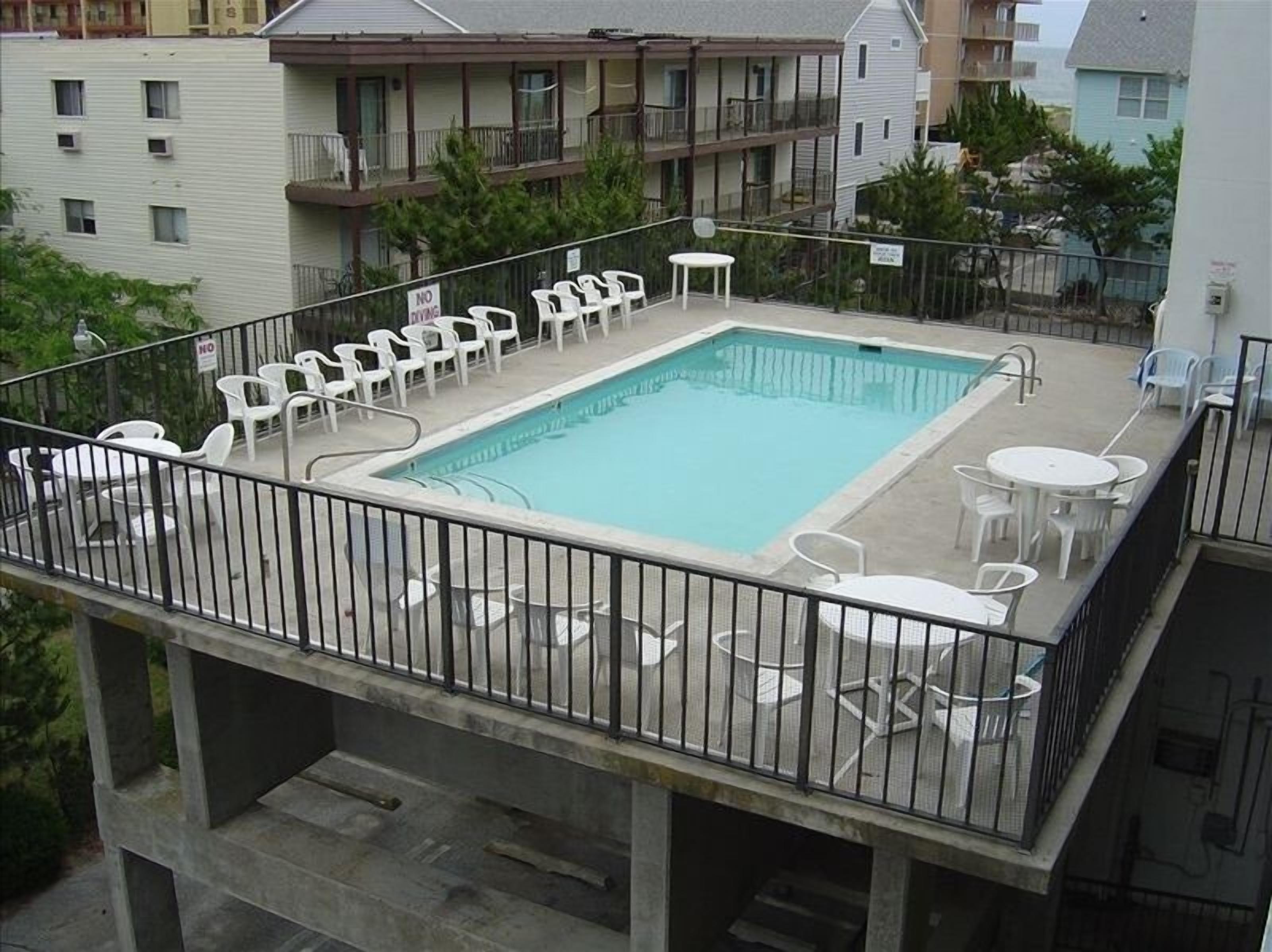 A rooftop pool with a square shape, surrounded by white plastic chairs and tables, and a metal railing.