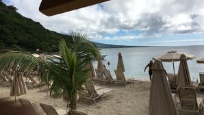 Een strand met ligbedden en parasols, een palmboom en uitzicht op zee en heuvels.