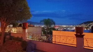 Vista nocturna desde un balcón con barandilla, un árbol y un paisaje urbano al fondo.