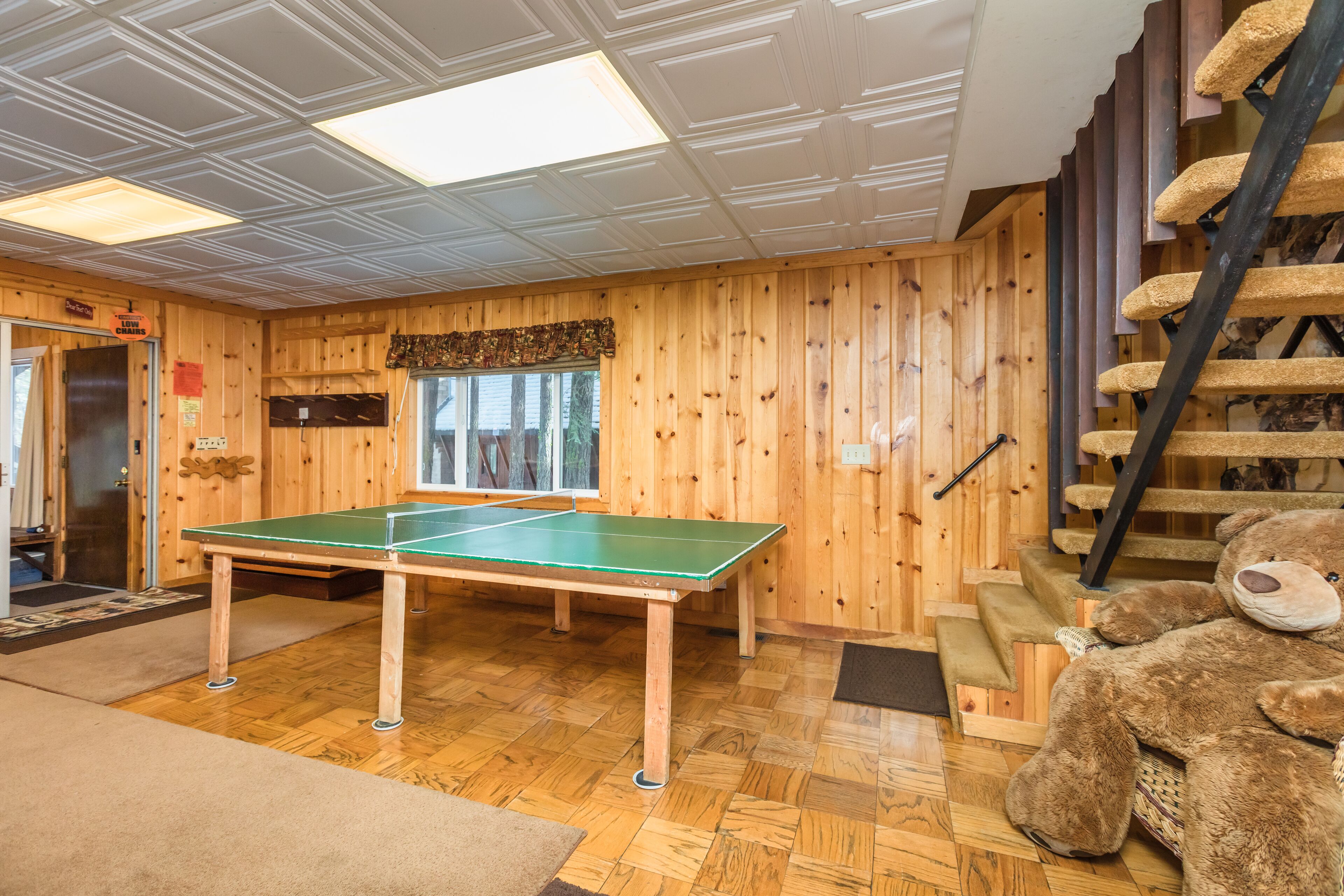 A room with a ping pong table, wooden walls, a large teddy bear, and a staircase.