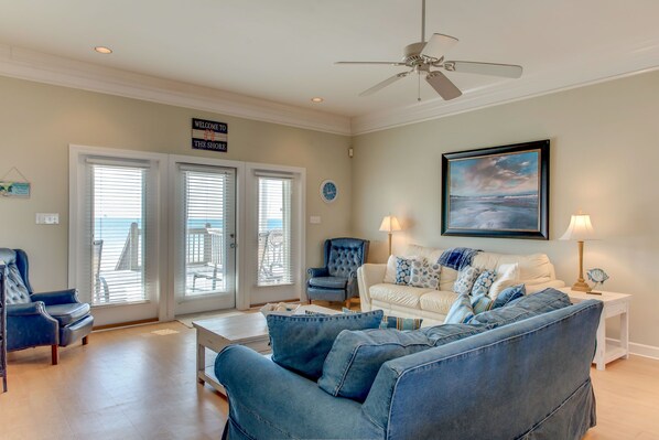 A living room with a white sofa, blue armchairs, and a coffee table. There is a ceiling fan, a framed painting, and a sign that reads "Welcome to the Shore."