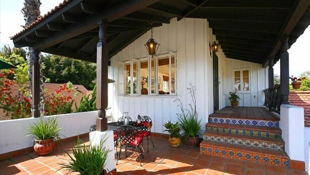A covered patio area with a table and chairs, potted plants, and a staircase leading to a white building.