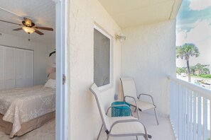A balcony with two chairs, a ceiling fan, and a view of palm trees and a road. - Beachside Bliss (Jacksonville Beach)