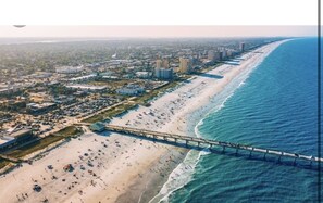 An aerial view of a beach with a long pier extending into the ocean, surrounded by buildings and a parking lot. - Beachside Bliss (Jacksonville Beach)