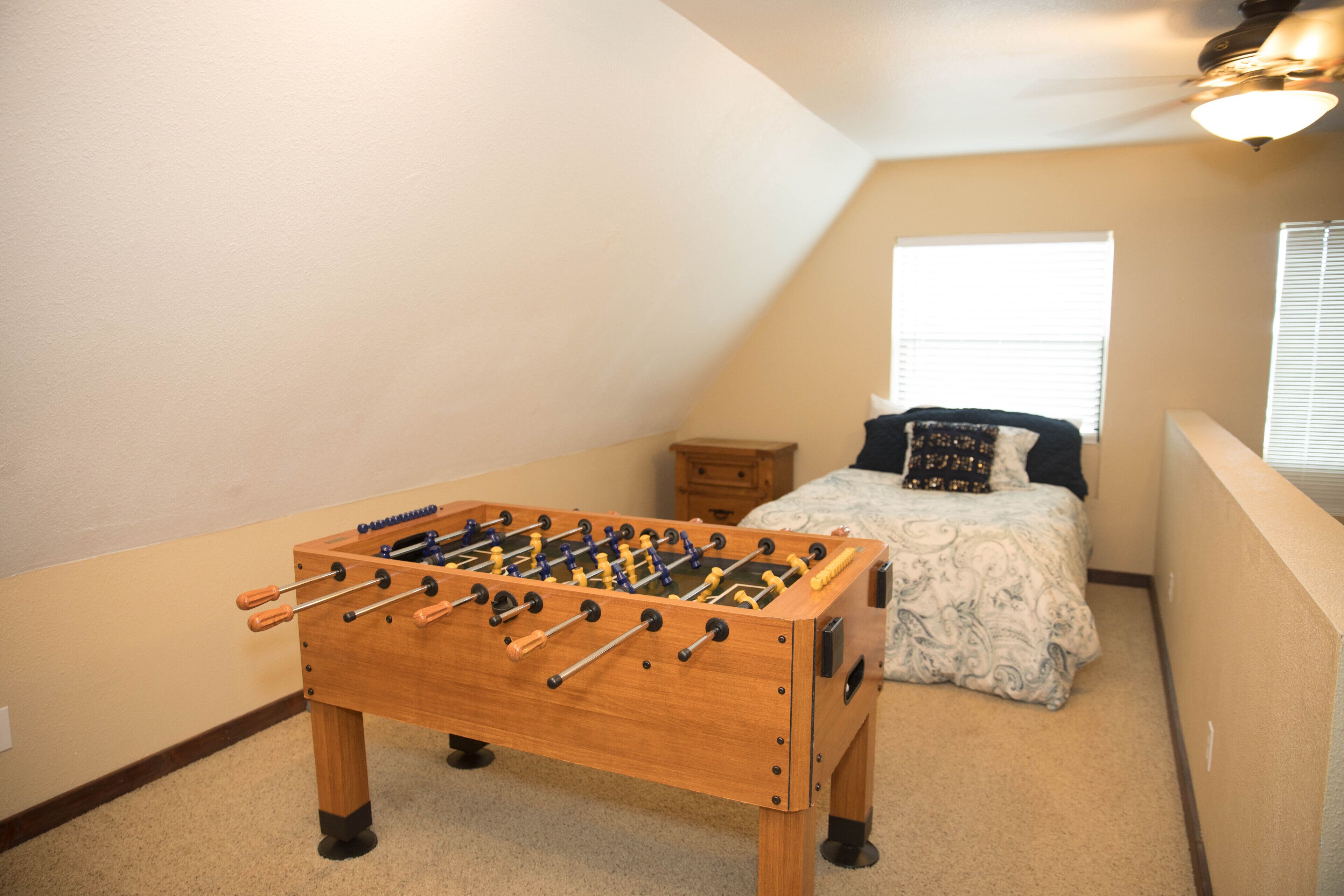 Een kamer met een tafelvoetbalspel, een bed met een decoratieve sprei, een nachtkastje en een raam met jaloezieën.