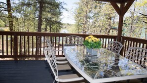 A glass-top table with white metal chairs on a wooden deck, surrounded by trees and a view of a lake. - SPECTACULAR MOUNTAIN BLUFF CABIN WITH LAKE VIEWS-CENTRAL AC! (Pinetop-Lakeside)