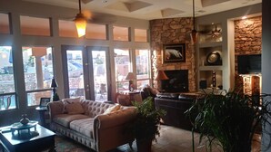 A spacious living room with a stone fireplace, leather sofa, and large windows overlooking a pool area. - THE RETREAT, an Executive Custom Home in Estrella Mtns! (Goodyear)