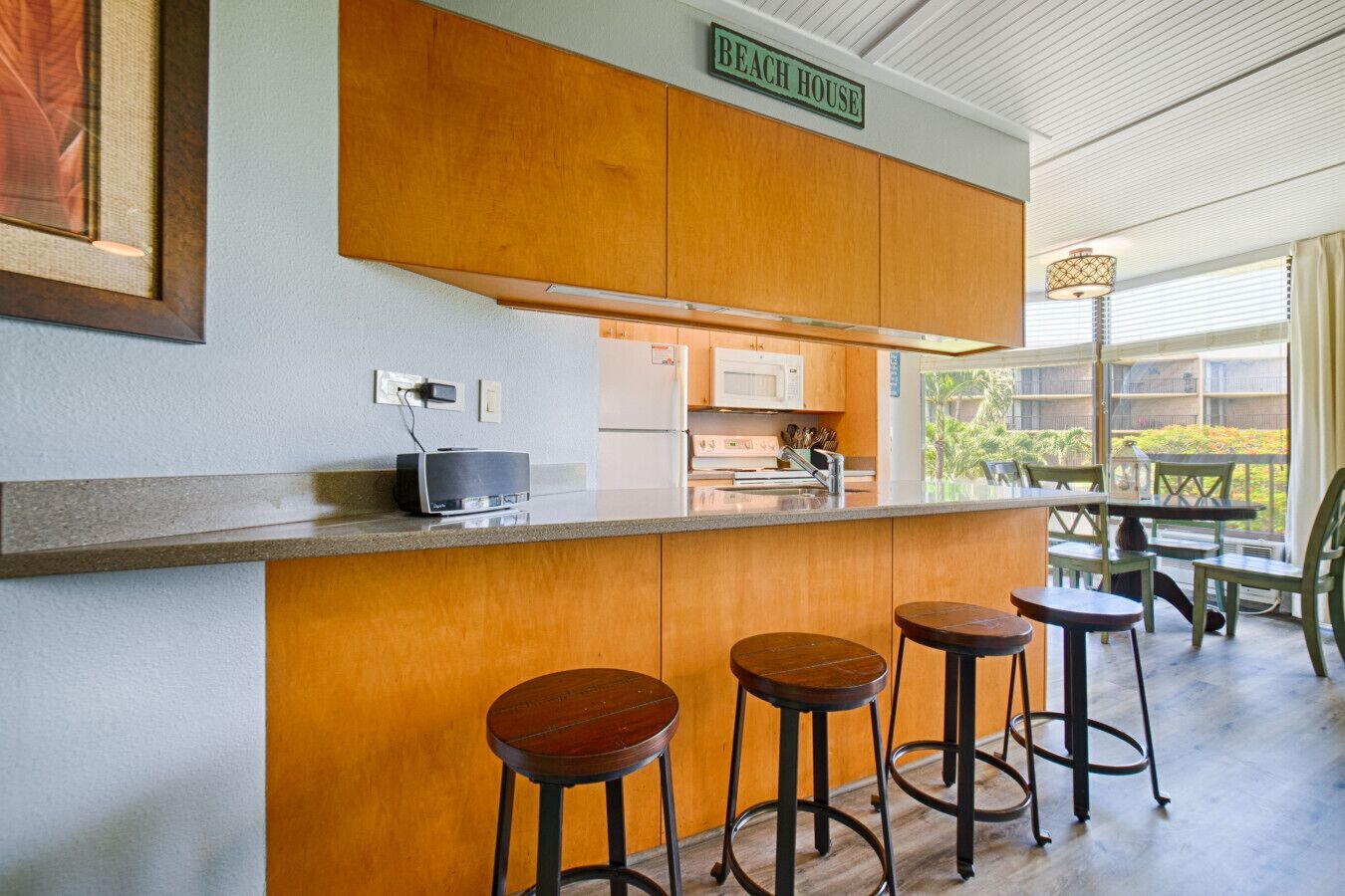 A kitchen with wooden cabinets, a microwave, and a coffee machine. There are three stools and a table with chairs. A sign above the counter reads "BEACH HOUSE".