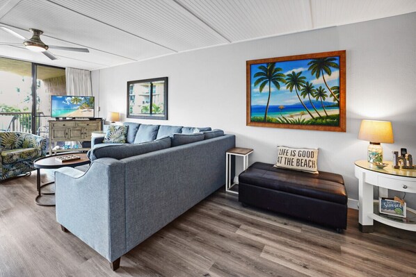 A modern living room with a grey sofa, a coffee table, a side table with a lamp, a framed painting of palm trees, and a sign that reads "LIFE IS GOOD AT THE BEACH".