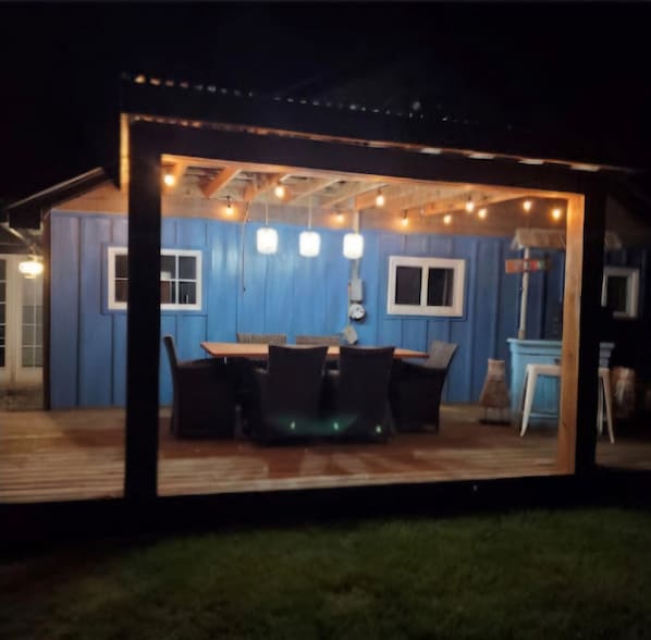 Un área de comedor al aire libre cubierta, con una pared azul, una terraza de madera y luces colgantes.