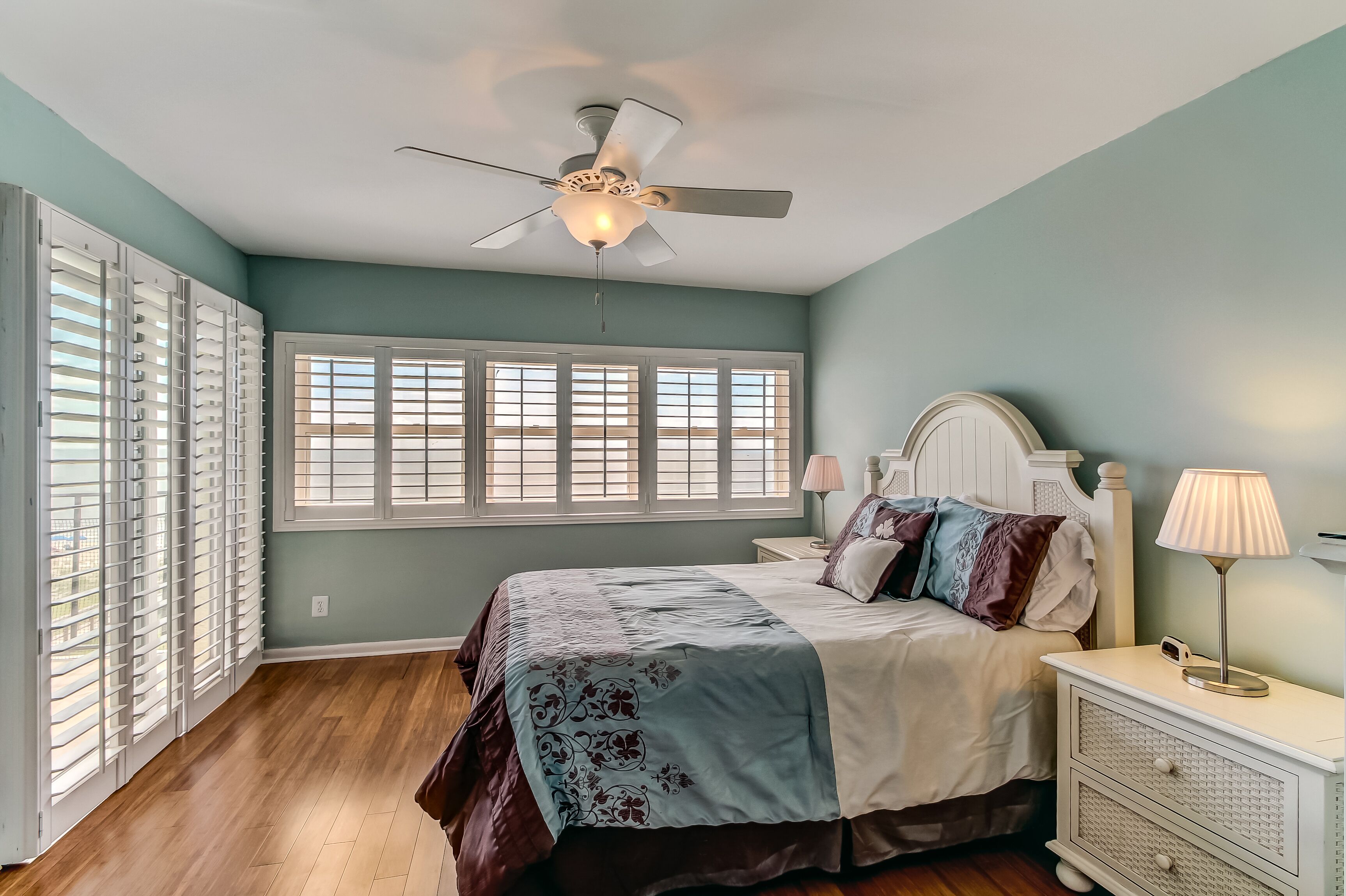 Une chambre à coucher comprenant un lit, une table de chevet, une lampe, un ventilateur de plafond et de grandes fenêtres équipées de stores.