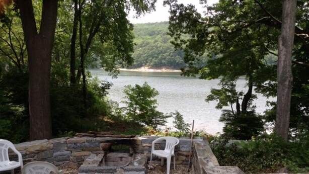 A stone fire pit with two white plastic chairs and a stone wall surrounding it, overlooking a lake with trees and a distant shoreline. - Waterfront! Charming 4BR Cottage on the Niantic River!  (Waterford)