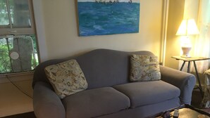 A living room with a grey sofa, patterned cushions, a painting of a beach scene, a standing fan, a lamp, and a small wooden table with a keyboard. - Waterfront! Charming 4BR Cottage on the Niantic River!  (Waterford)