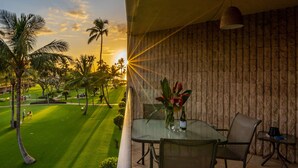 Ein Balkon mit Tisch und Stühlen, der auf eine gepflegte Rasenfläche und Palmen mit Sonnenuntergang im Hintergrund blickt.
