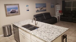 Una cocina con encimera de granito, pileta de acero inoxidable y muebles blancos. Hay un bote de basura, un refrigerador y un ventilador de techo.