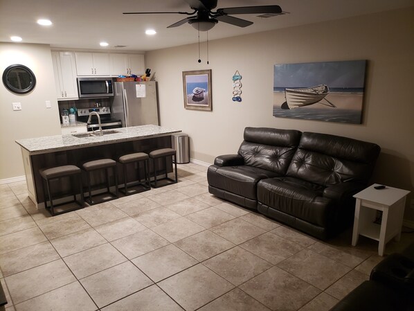 A modern kitchen with a central island, a black leather sofa, and a painting of a boat on the wall. - Beautiful Pool - 2 BR/2 BA - 1 Mile to Beach (Port Aransas)