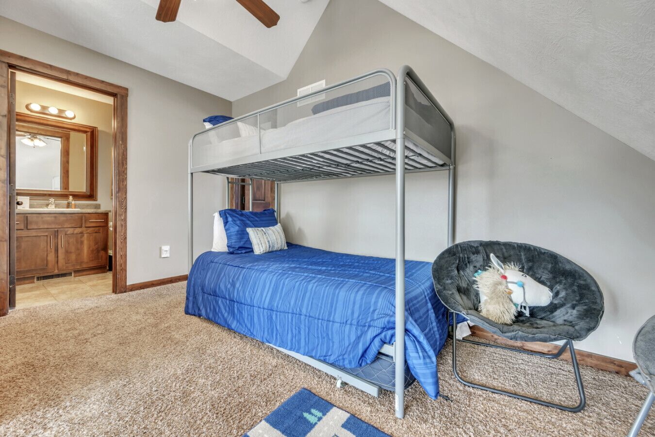 A bunk bed with a blue bedspread, a chair with a plush toy, and a bathroom with a vanity and mirror.
