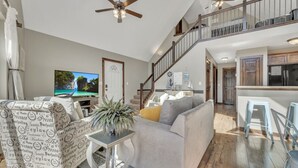 Ein großzügiges Wohnzimmer mit grauem Sofa, gemustertem Sessel und Fernseher. Eine Treppe führt zu einer oberen Ebene, und im Hintergrund ist eine Küche zu sehen.
