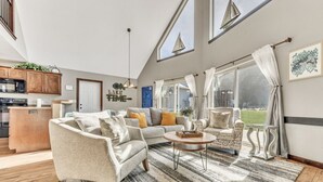 Ein geräumiges Wohnzimmer mit einer Couch, Sesseln und einem Couchtisch. Es gibt eine Küchenzeile mit Holzschränken und einer Mikrowelle. Große Fenster mit Vorhängen und Bootsdekorationen sind zu sehen.