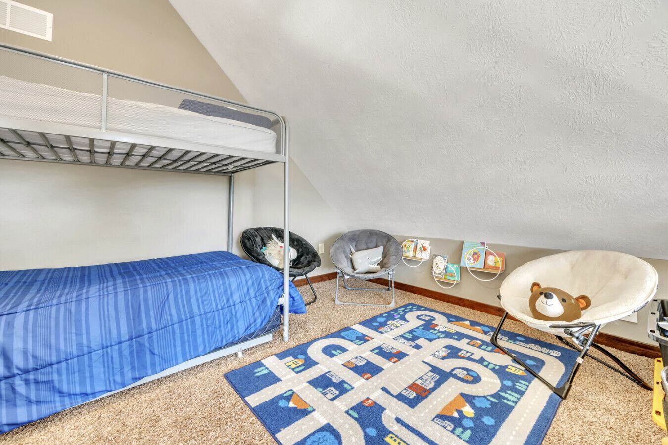 A dormitory room with bunk beds, a desk, a chair, a rug, and wall decorations.