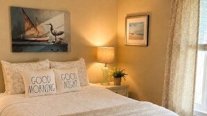 Un dormitorio con cama, almohadas, lámpara de noche, un cuadro, ventana con cortinas y una mesita con una planta.