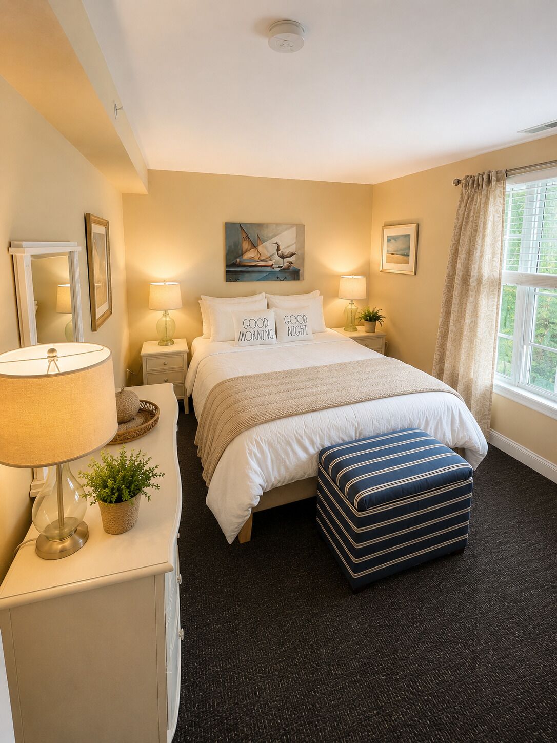 A bedroom with a bed, bedside lamps, a nightstand, a window with curtains, and a painting on the wall.