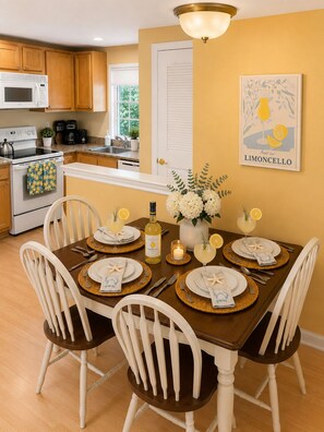 A dining area with a table set for four, a kitchen in the background, and a framed artwork on the wall. - Goose Rocks Beach!!  Sunny bright condo. (Kennebunkport)