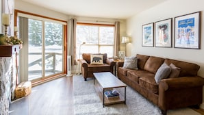 A cozy living room with a brown leather sofa, a wooden coffee table, and a large window with a view of snow-covered trees. - SKI IN/OUT, close to gondola and village - 1 bd, Hot Tubs/pool (Whistler)