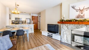 A modern kitchen with white cabinets, a dining area with a white table and chairs, a television on a white stand, and a stone fireplace with a deer painting above it. - SKI IN/OUT, close to gondola and village - 1 bd, Hot Tubs/pool (Whistler)