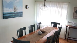 Comedor con mesa de madera, seis sillas, un cuadro de un barco en la pared, ventilador de techo y ventana con cortinas traslúcidas.