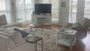 Una sala de estar con un sofá blanco, una mesa de centro, un televisor sobre un mueble bajo y un ventilador de techo.