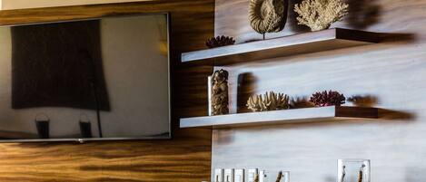 A modern living room with a flat-screen TV mounted on a wooden wall, decorative shelves with shells and a small statue, and a glass cabinet with rope handles.