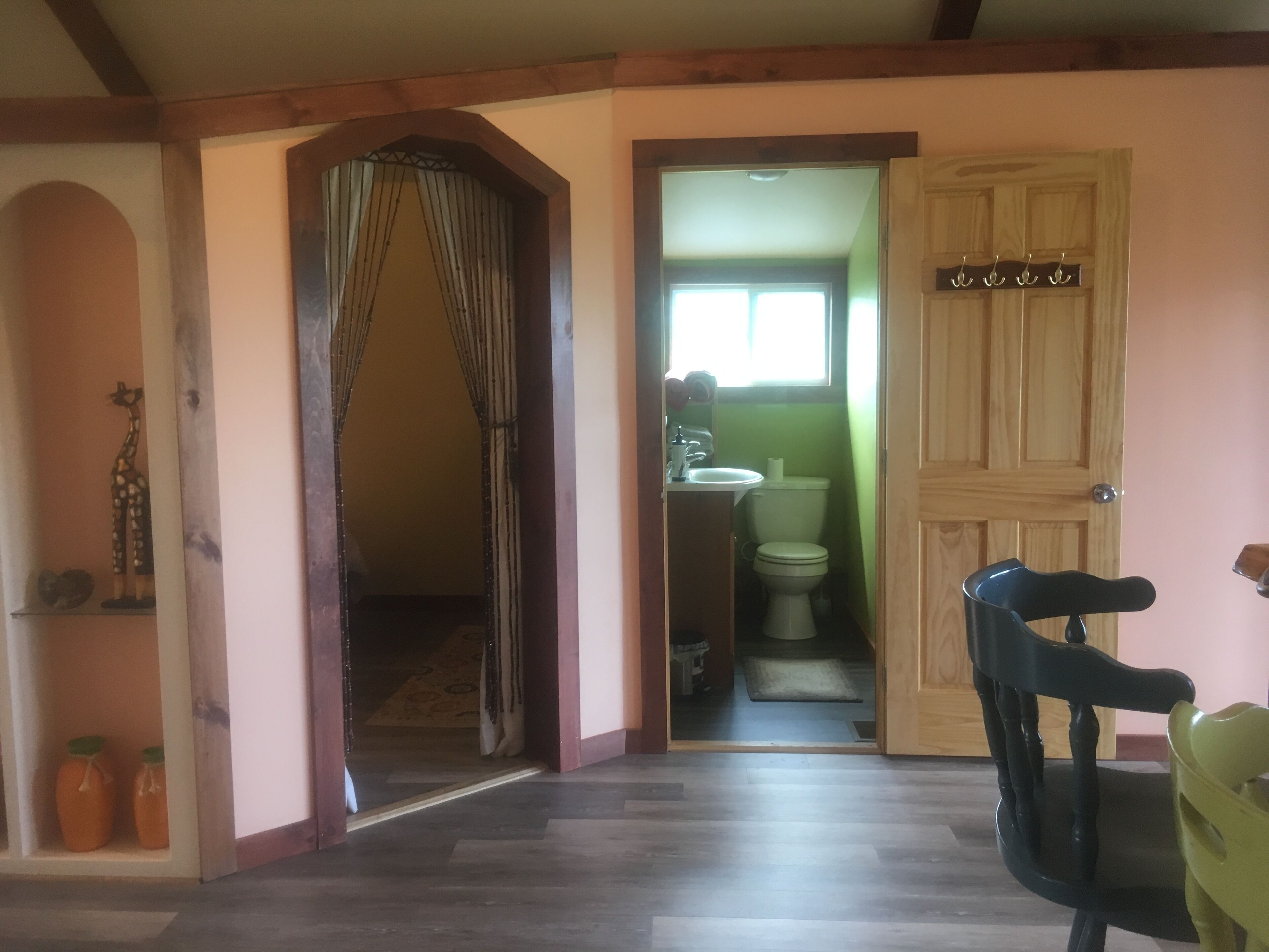 A hallway with a wooden door, a small bathroom with a toilet and sink, and a chair in the foreground.