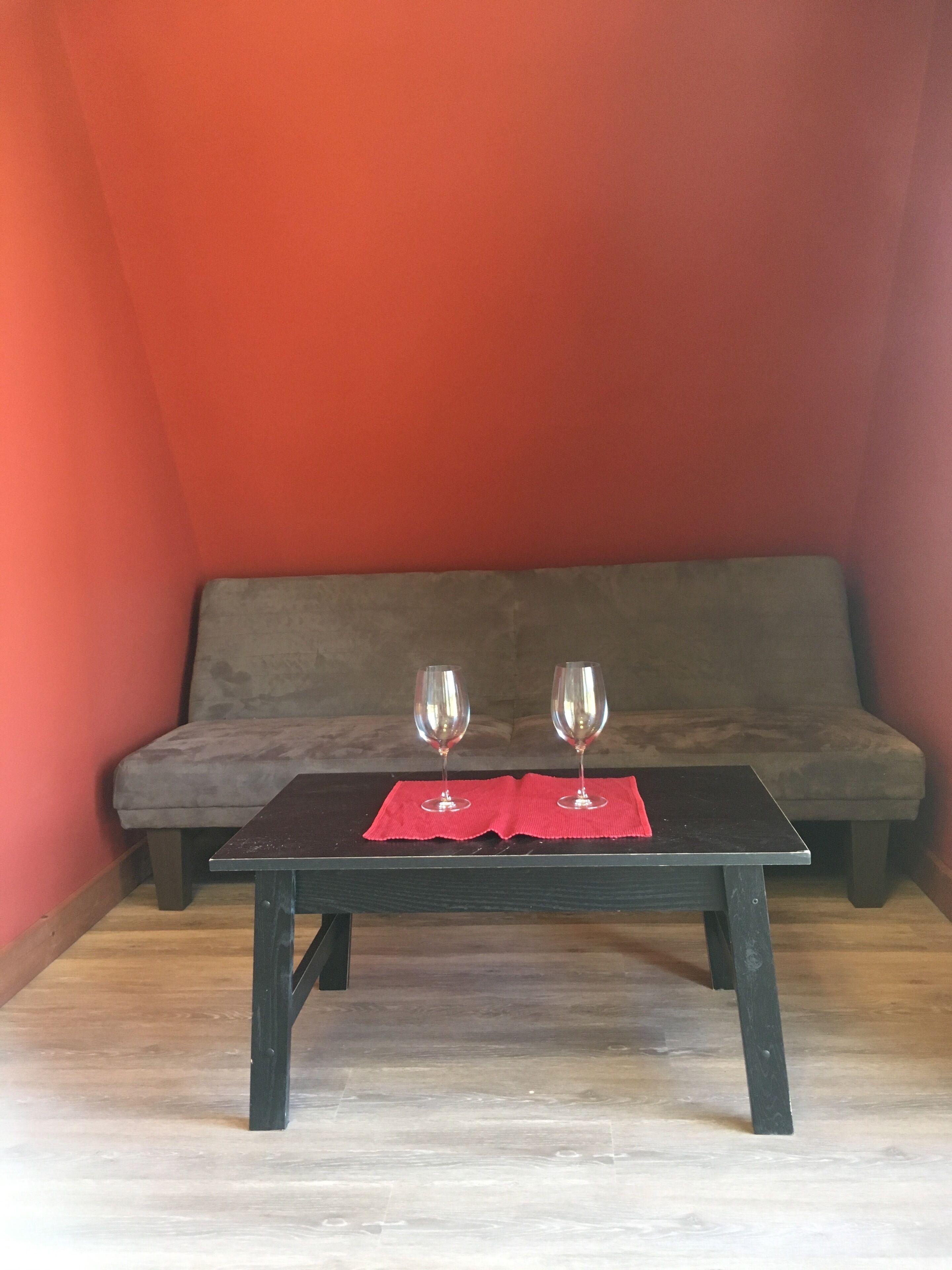 A small room with a dark wooden table, two wine glasses on a red cloth, and a grey sofa against a red wall.