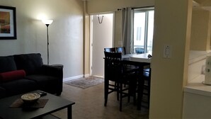 Una sala de estar con un sofá negro, una mesa de centro y un cuadro en la pared. Hay un comedor con una mesa y sillas, y una ventana con cortinas.