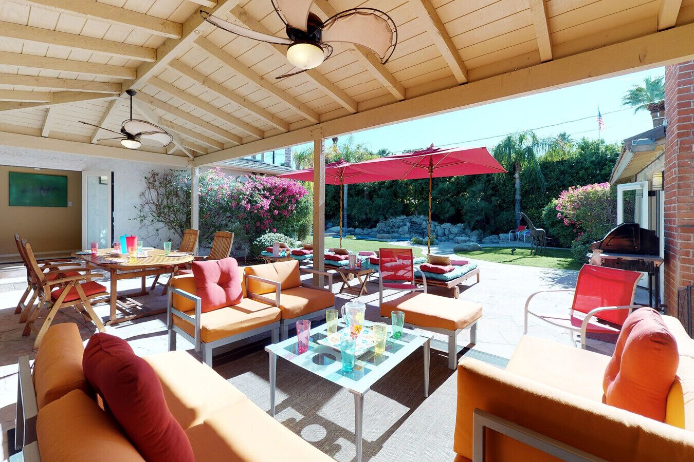 An outdoor patio with wooden furniture, red umbrellas, and a view of the backyard.