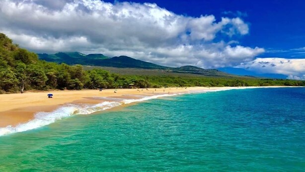 Une plage à l’eau turquoise et limpide, bordée d’un rivage sablonneux, avec en arrière-plan des collines verdoyantes sous un ciel partiellement nuageux.