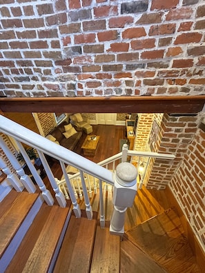 A staircase with a wooden handrail leading to a room with a brick wall, a wooden floor, and a seating area with cushions. - Stylish Escape in Historic Warehouse District (New Orleans)