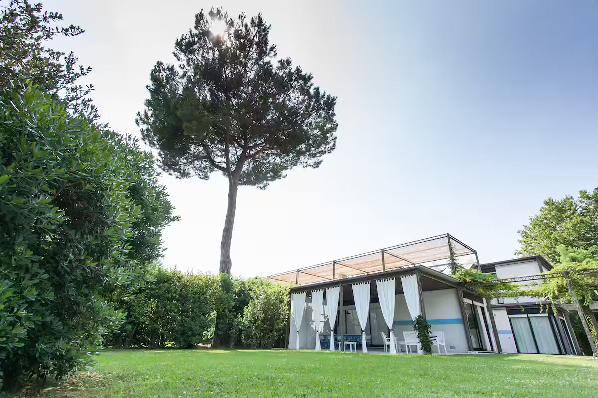 A modern building with a covered outdoor area, surrounded by greenery and a large tree.