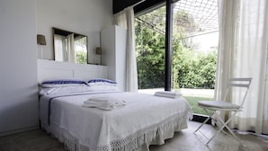 A modern bedroom with a large bed, a chair, and a view of greenery outside. - Architectural/Minimal Villa in Forte dei Marmi, Roma Imperiale (Forte dei Marmi)