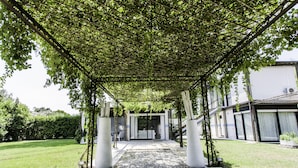 A covered walkway leading to a building with a modern entrance, surrounded by greenery and a gravel path. - Architectural/Minimal Villa in Forte dei Marmi, Roma Imperiale (Forte dei Marmi)