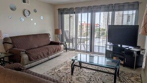A living room with a brown sofa, a glass coffee table, a TV, and a view of a balcony with outdoor furniture. - Right on the Beach,  Ocean View, Modern 2BR/2B, 2 balconies (Cocoa Beach)