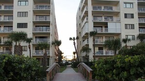 A pathway leading to a multi-story residential building with balconies, surrounded by greenery and palm trees. - Right on the Beach,  Ocean View, Modern 2BR/2B, 2 balconies (Cocoa Beach)