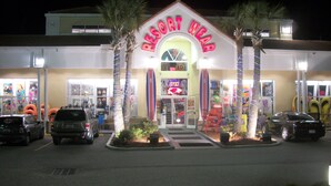 A resort wear store with a neon sign, palm trees, and parked cars. - Beautiful Efficiency Condo Across The Street from Beach. Great Pool & Deck Area. (North Myrtle Beach)